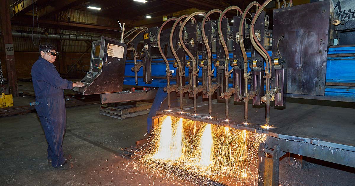 Worker controlling a multi-torch flame cutting machine, with bright sparks flying as thick metal is being cut.